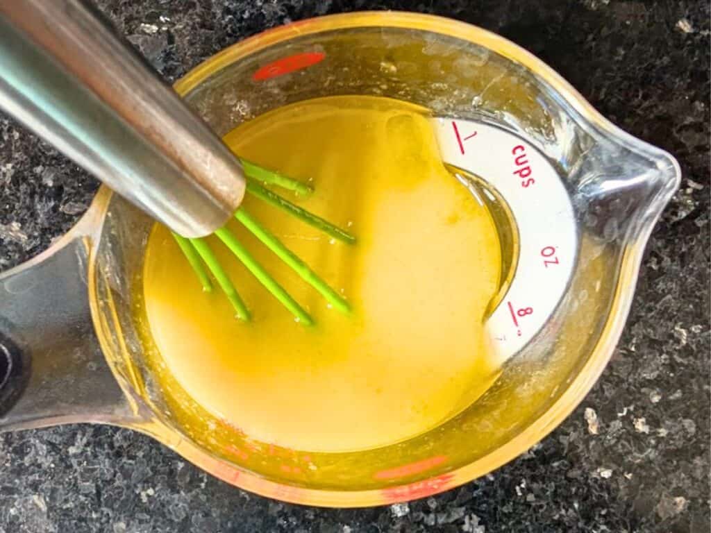 Eggs and oil whisked together in a glass measuring cup with a green whisk.