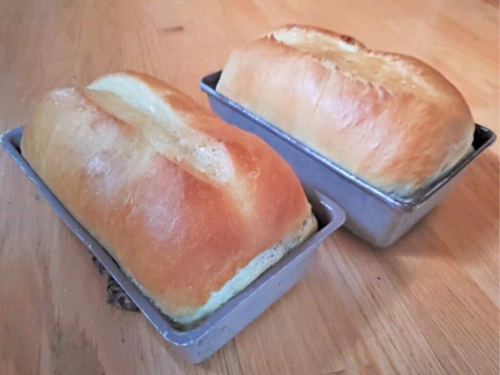 Two freshly baked loaves of bread in rectangular metal pans on a wooden surface.