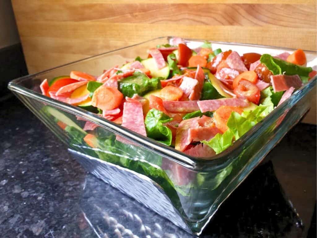 A glass bowl filled with a salad containing lettuce, sliced carrots, apple pieces, and strips of deli meat, tossed in a homemade dijon vinaigrette and placed on a dark countertop.