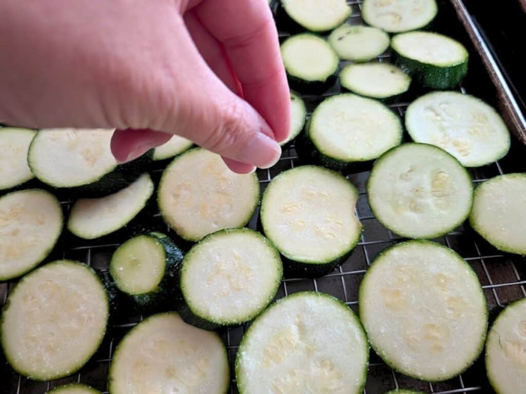 A hand sprinkles seasoning over raw zucchini slices arranged on a wire rack for preparation.