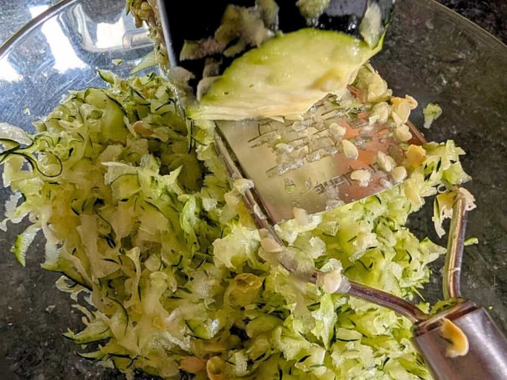 A close-up of a zucchini being grated with a box grater over a glass bowl, with shredded zucchini collecting below.