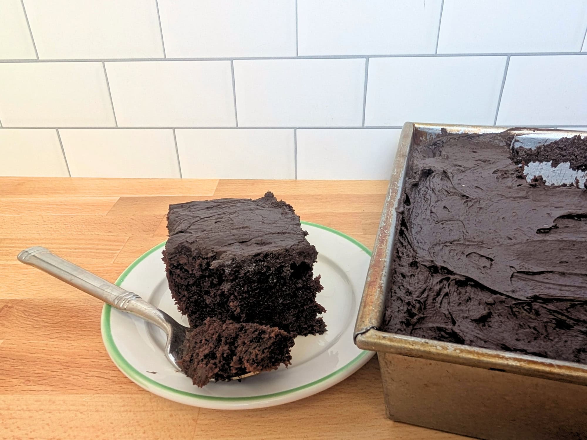 A square slice of chocolate cake on a plate with a fork, next to a rectangular pan filled with frosted chocolate cake.