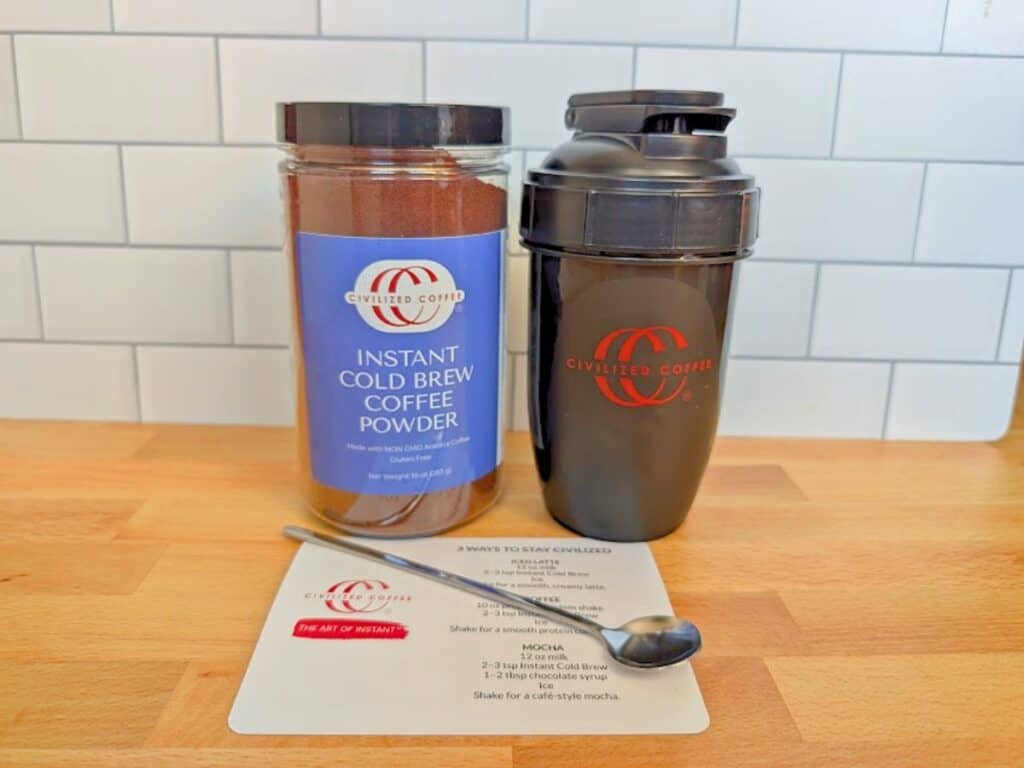 A jar of Civilized Coffee instant cold brew powder, a shaker bottle, a metal spoon, and a recipe card on a wooden surface.