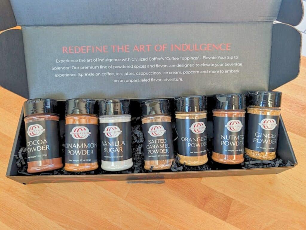 A gift box containing seven labeled jars of Civilized Coffee drink toppings, including cocoa, cinnamon, vanilla sugar, salted caramel, orange zest, nutmeg, and ginger powder.
