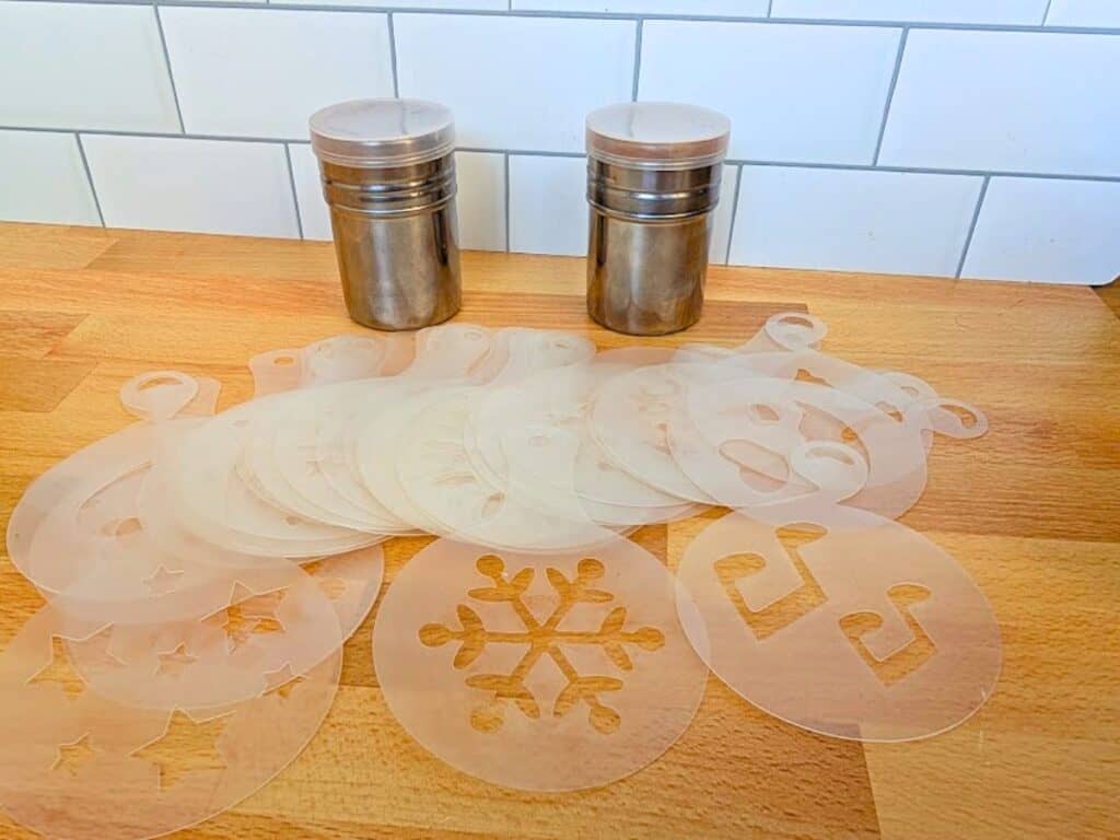 A stack of translucent plastic coffee stencils with various patterns, including a snowflake and stars, next to two stainless steel shakers on a wooden counter.