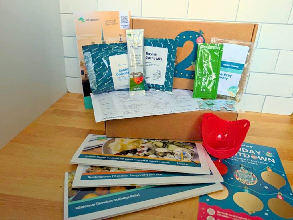 An Eat2explore meal kit box with spice packets, recipe cards, a red egg separator, and instructional pamphlets on a wooden surface.