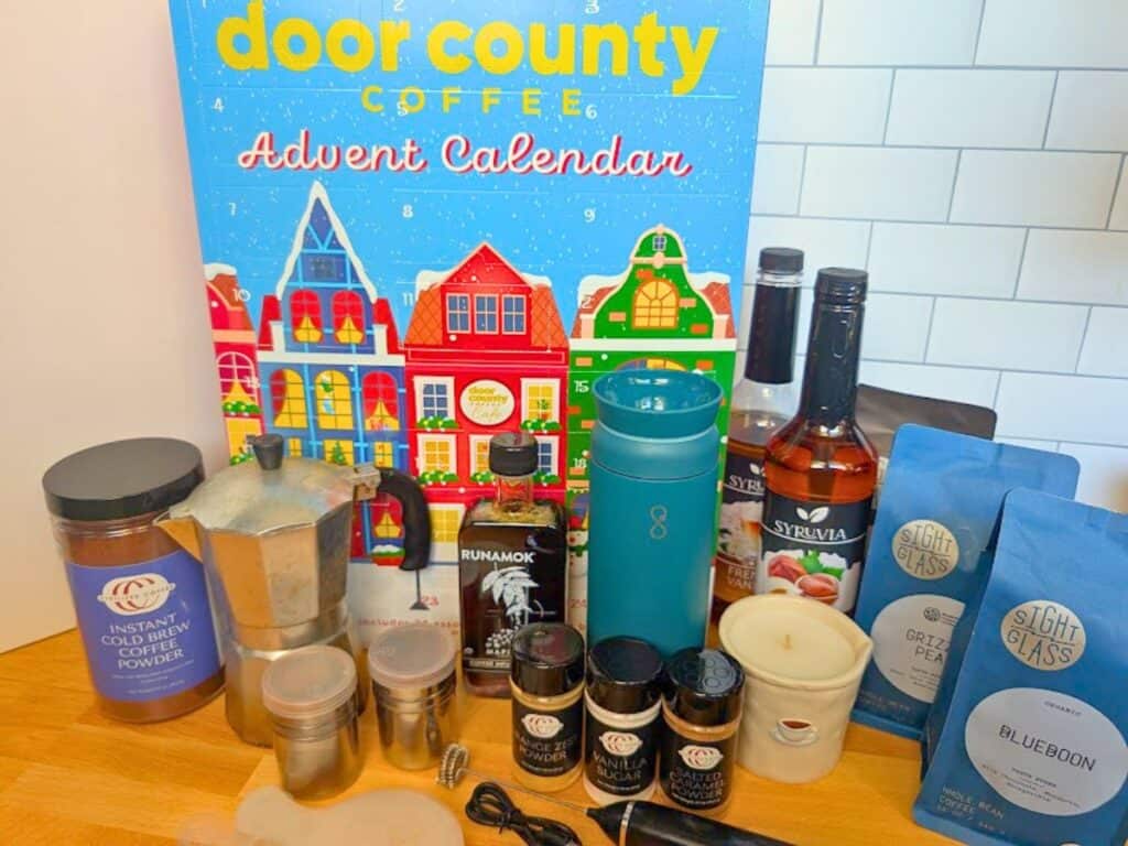 A Door County Coffee advent calendar surrounded by coffee accessories, including a moka pot, syrups, spices, a travel mug, and two bags of Sightglass coffee beans.