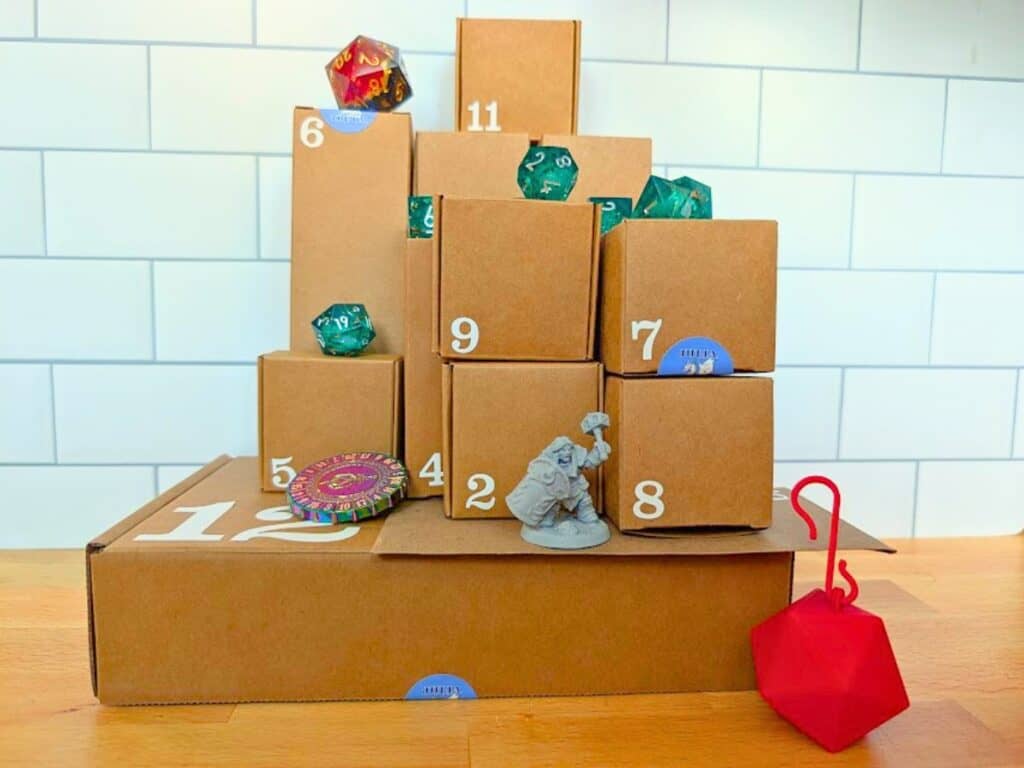 A tabletop gaming advent calendar made of stacked cardboard boxes numbered 2 through 11, accented with scattered polyhedral dice, a miniature figure, and a circular token.
