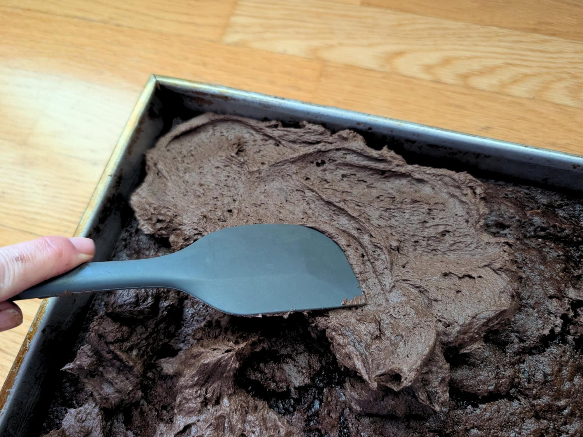 A hand uses a gray spatula to spread chocolate frosting over a sheet cake in a metal pan.