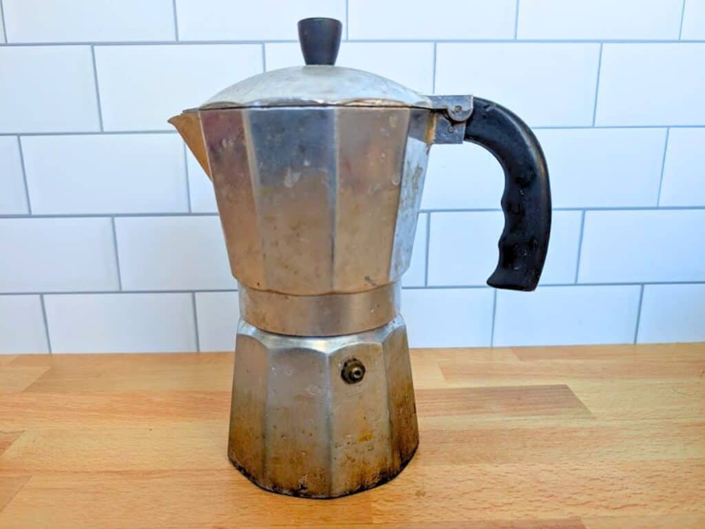 A metal Moka pot with a black handle and lid knob sitting on a wooden countertop.