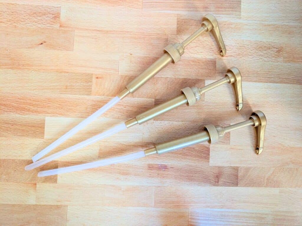 Three gold-colored bottle pumps with clear plastic dip tubes lying on a wooden surface.