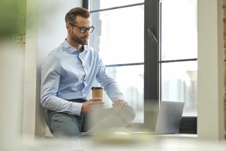 Elegant forretningsmand med kop kaffe og laptop ved vinduet i en moderne kontorindretning.