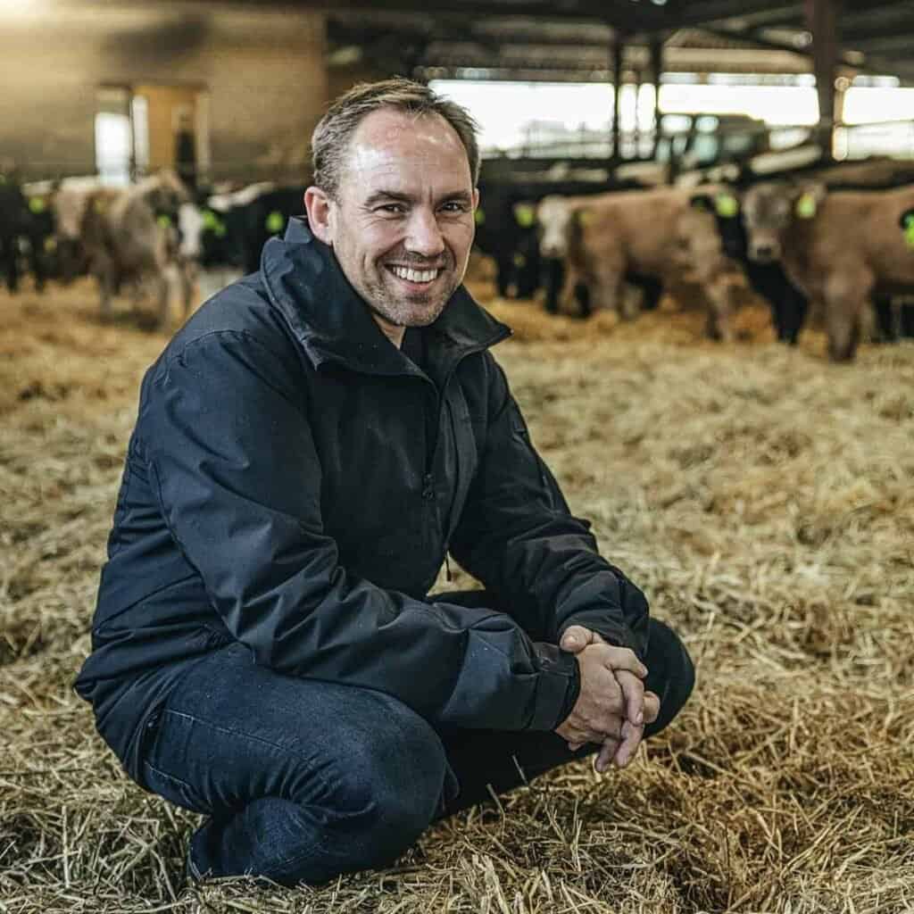 1. Billedet viser en smilende mand på en gård med kvæg i en staldmiljø.