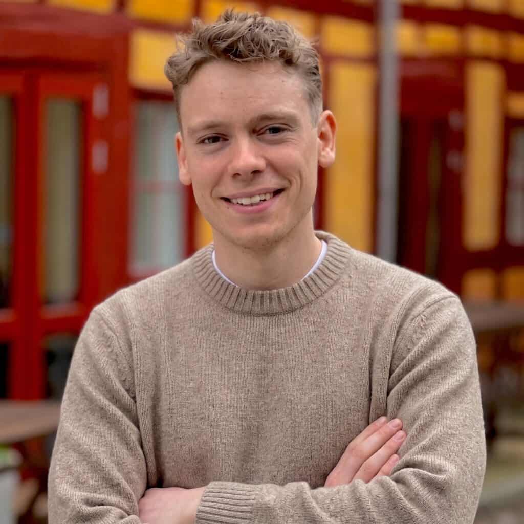 Mand smilende person i beige trøje udendørs med farverige bygninger i baggrunden.