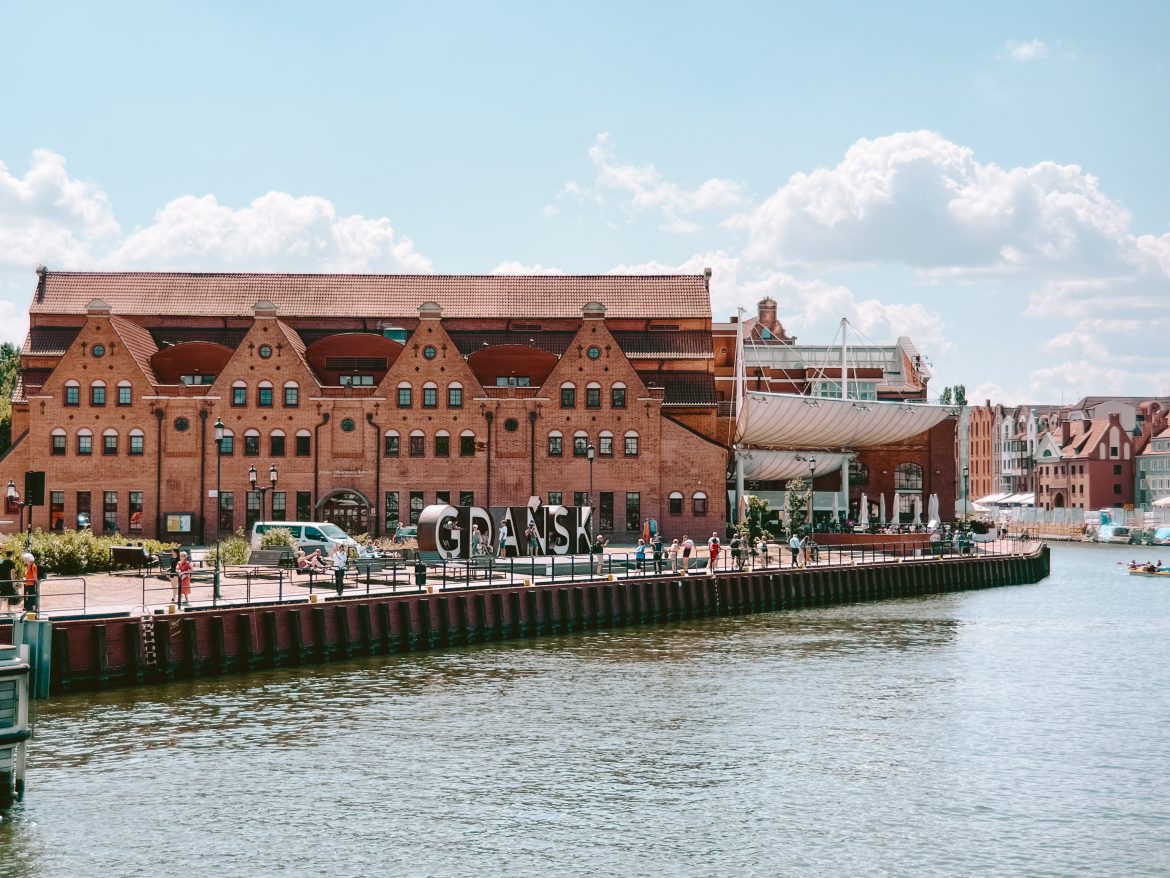 A brick building with arches stands majestically near the waterfront in Gdańsk, Poland, as people leisurely stroll along the promenade, savoring a delightful weekend in Gdańsk.