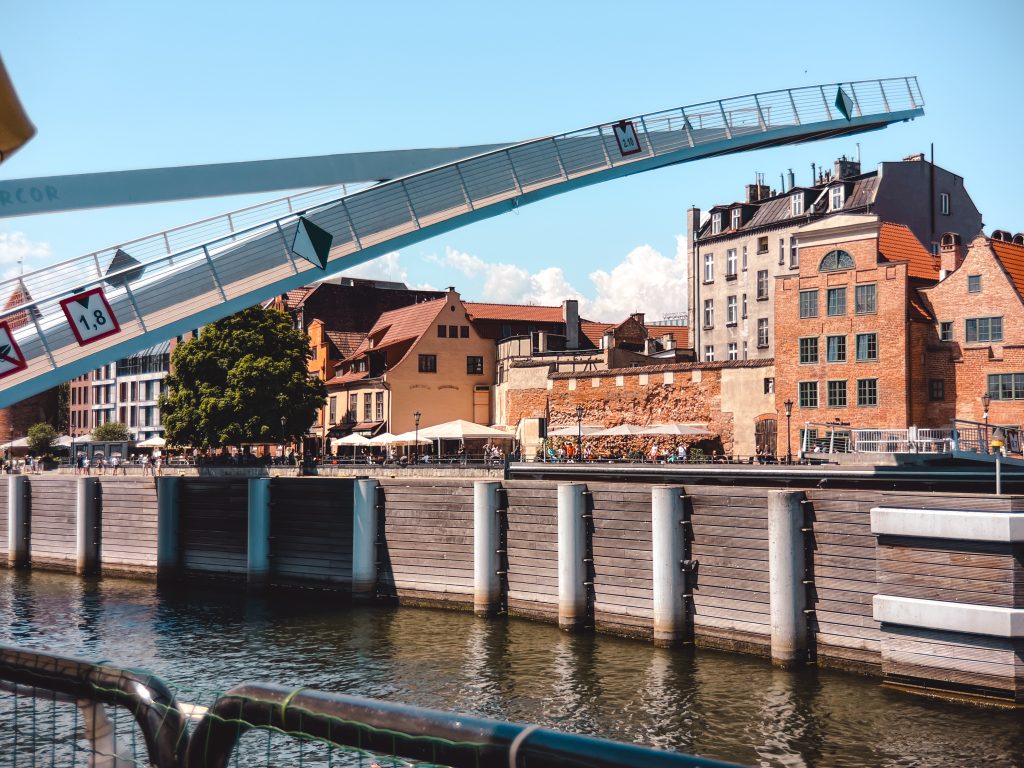 A drawbridge is raised over a river near old brick buildings and trees in Gdansk. The sky is clear and sunny.