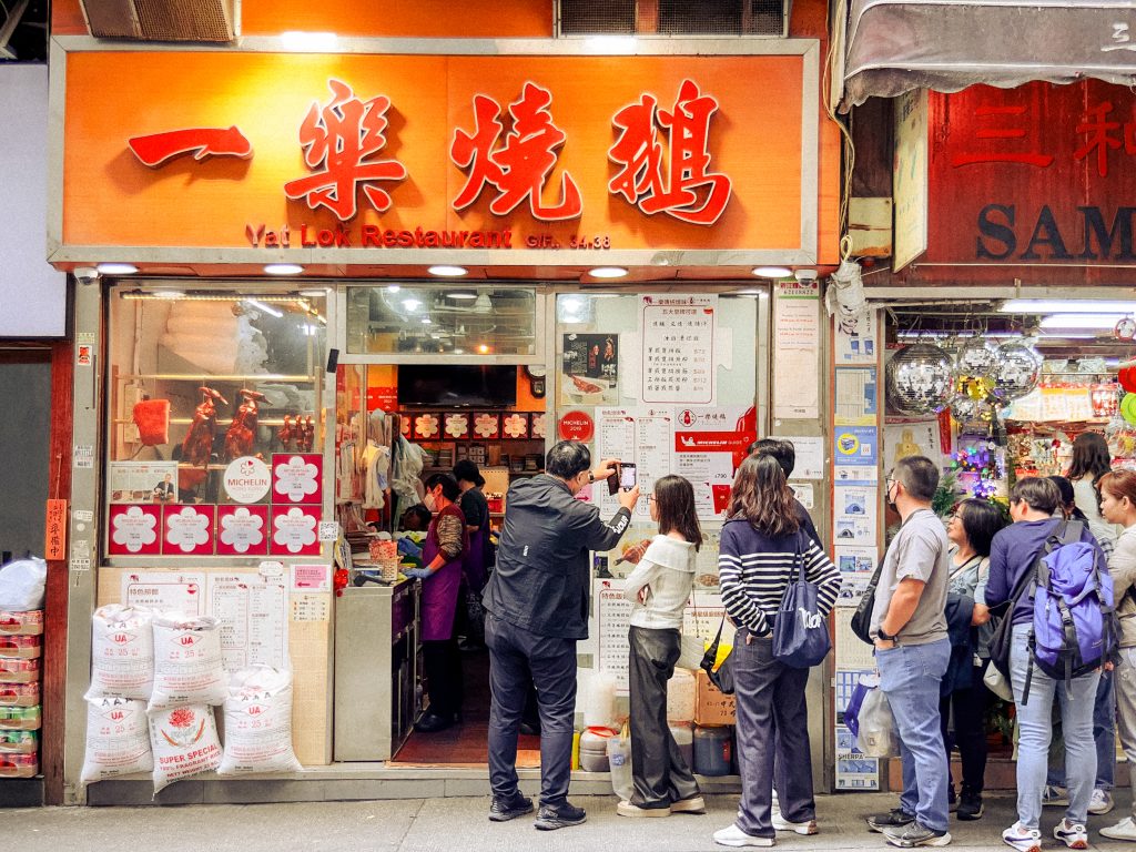 People wait in line outside Yat Lok Restaurant on a bustling street waiting for some of the best food in Hong Kong.