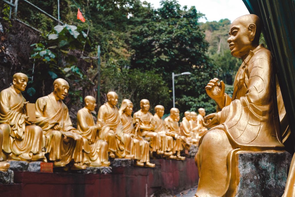 A row of golden monk statues, each in a seated position, lines a stone pathway surrounded by lush greenery.