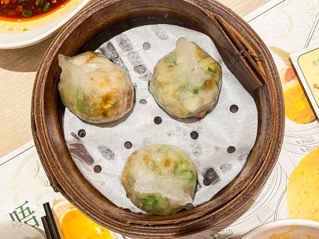 Three steamed dumplings rest in a bamboo basket on a paper liner, surrounded by dipping sauces on a table.
