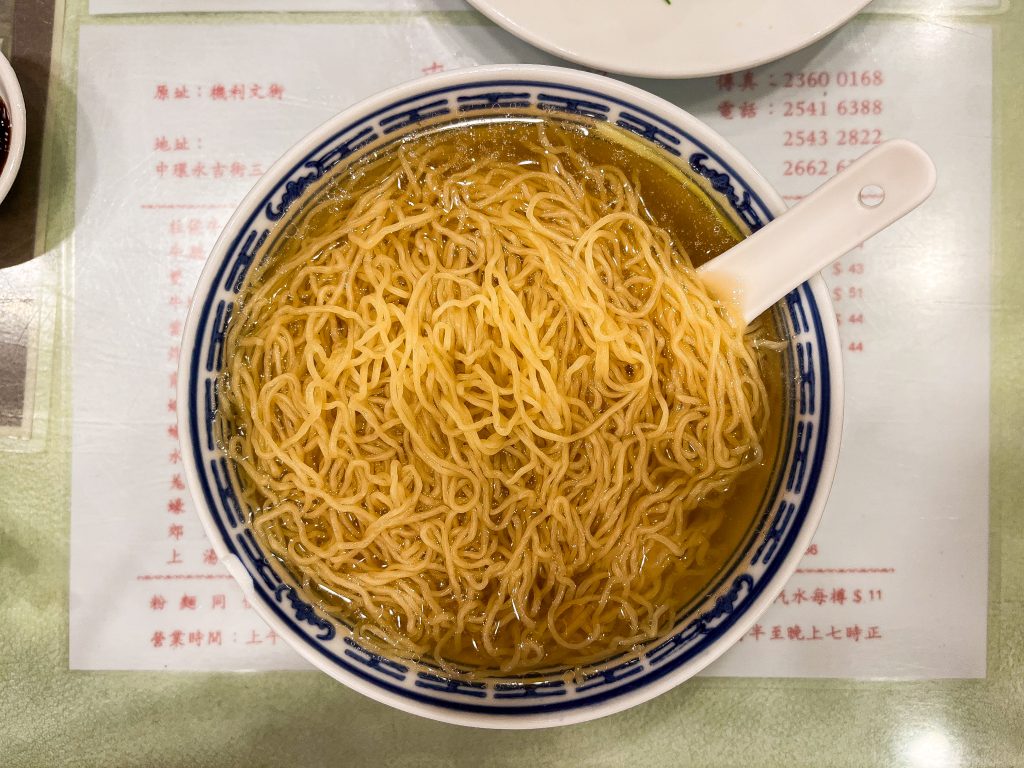 A bowl of yellow noodles in clear broth, served with a white soup spoon on a patterned bowl, sits invitingly on a menu-covered surface..