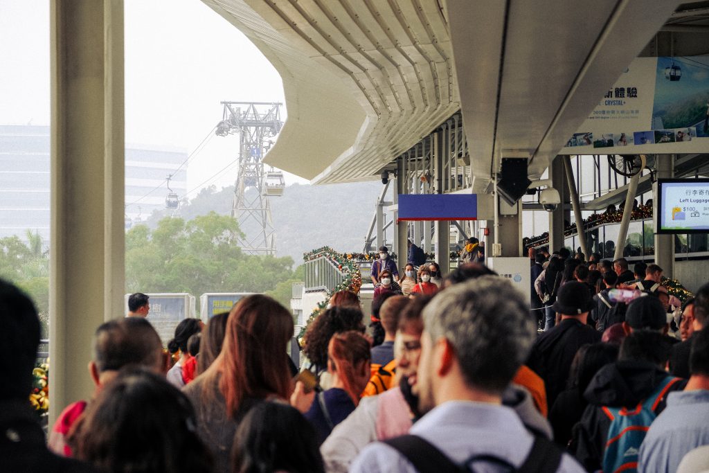 Crowds of people queue, waiting to board cable cars which can be seen disappearing into the Hong Kong greenery behind.