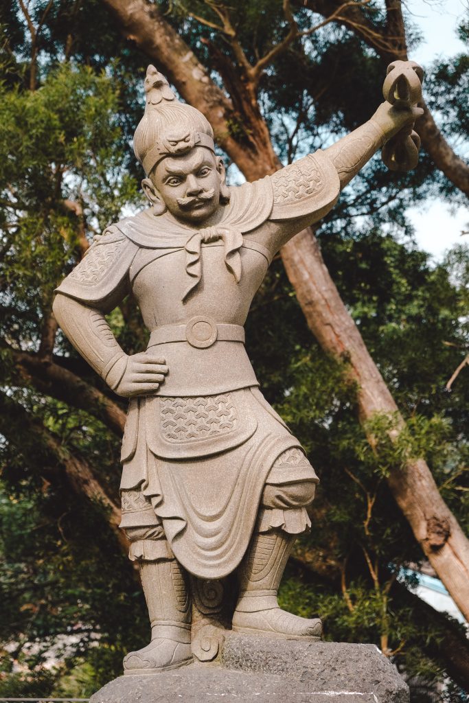 A stone statue of a warrior in ornate armour stands with one arm raised, framed by leafy trees.