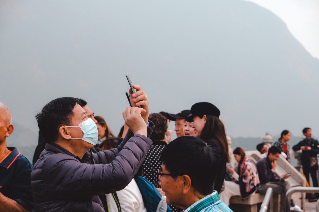 A crowd of people, some wearing masks, stands closely as a man takes a photo with his phone in an outdoor setting in Hong Kong.