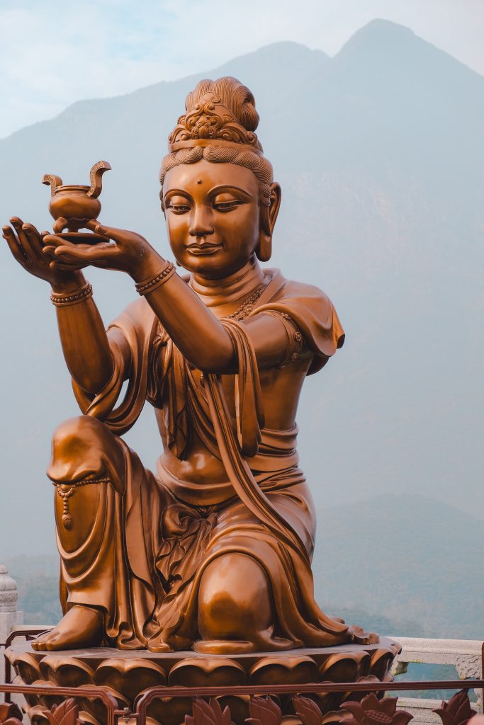 A bronze statue of a seated figure holding a small vessel, set against a stunning mountain backdrop.