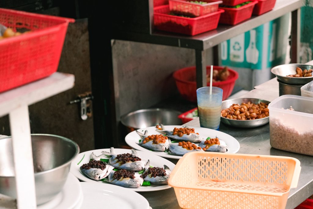 Plates of prepared fish with toppings on a kitchen counter, surrounded by baskets and a drink.