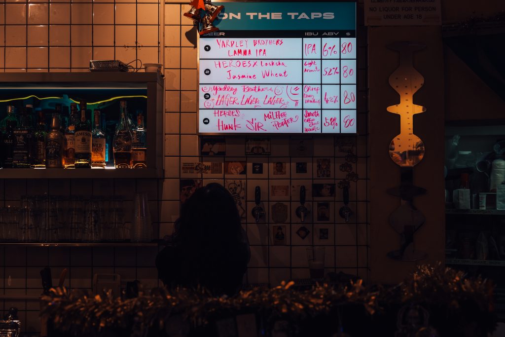 Bar interior with a person standing at a counter, illuminated beer menu on display above various liquor bottles.