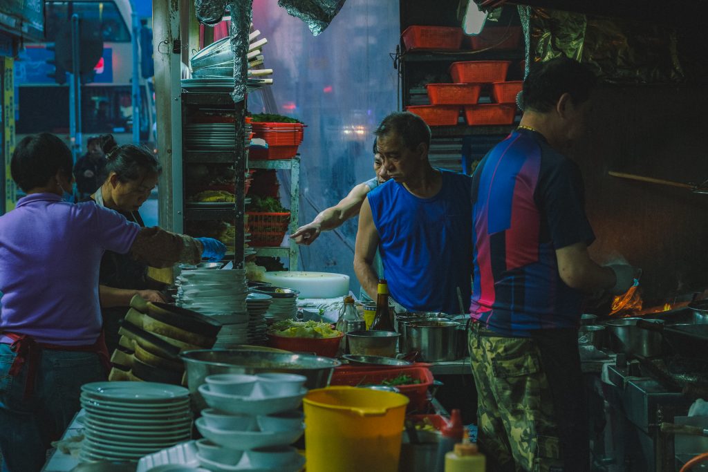 People cooking and preparing dishes in a busy outdoor kitchen with stacks of plates and various containers.