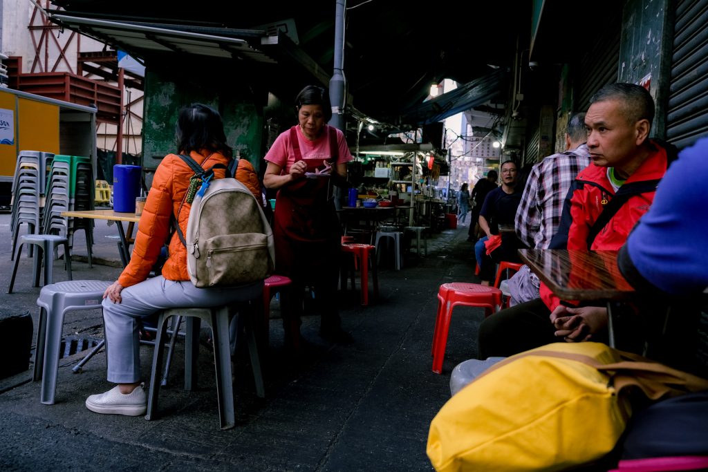 People seated at a street food stall in an alley, with a vendor serving a customer.