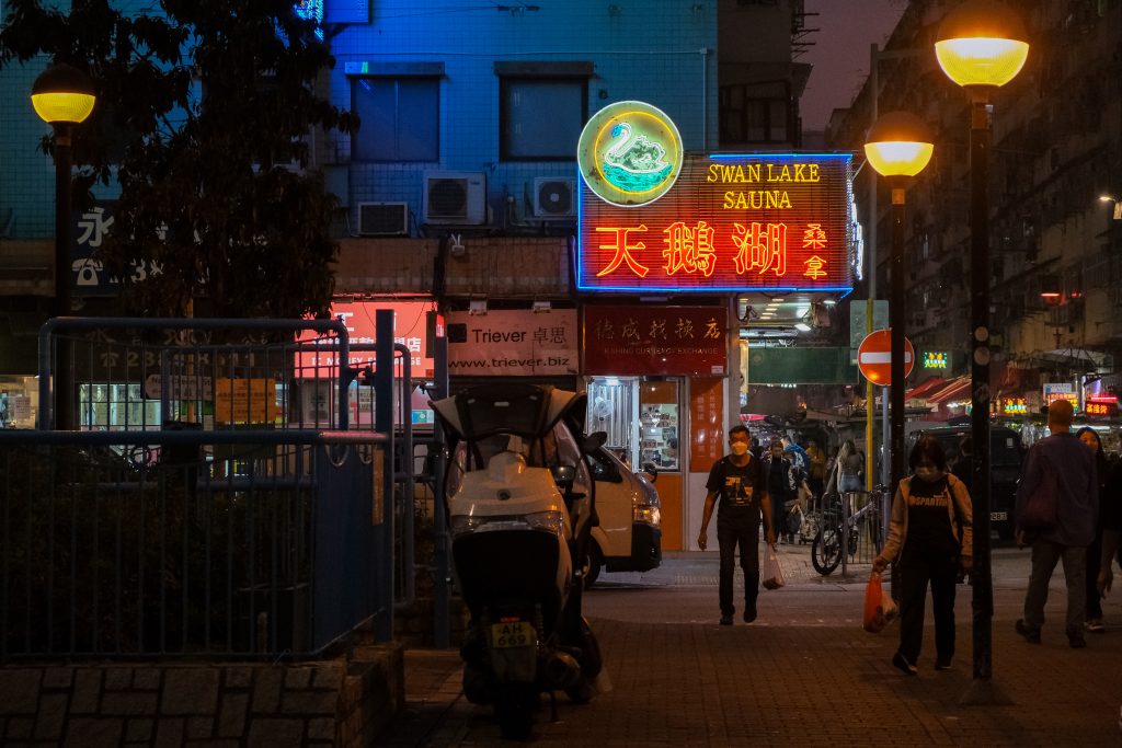 Night street with neon sign for Swan Lake Sauna, people walking, and a parked motorbike nearby.