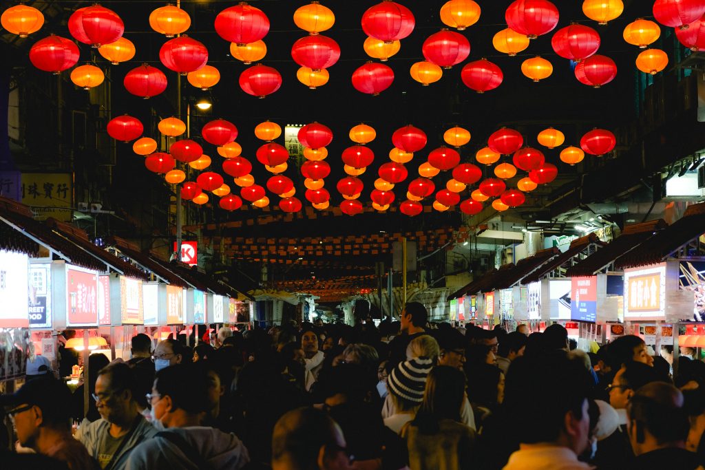 A weekend in Hong Kong comes alive at the crowded night market, where a canopy of red and orange lanterns glows overhead. Brightly lit stalls line both sides of the street, bustling with people exploring vibrant wares and tantalizing aromas.