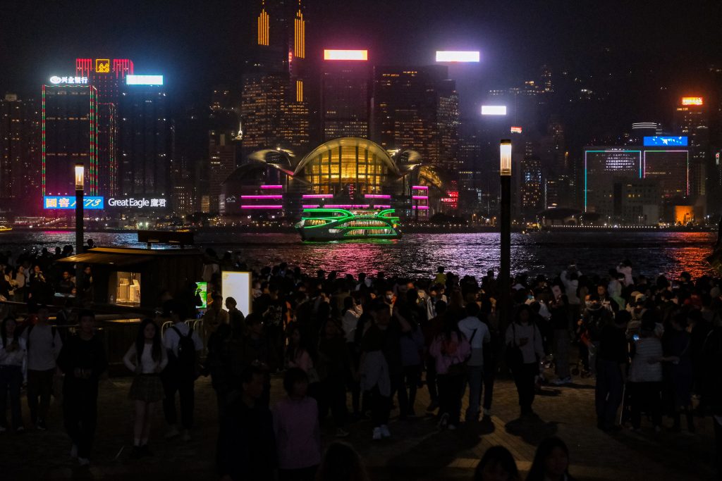 Nighttime cityscape with a crowd gathered along a waterfront, enjoying a weekend in Hong Kong. Brightly lit buildings and the iconic dome are visible in the background across the water, creating an enchanting urban glow.
