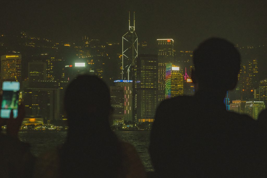 Silhouettes of people looking at Hong Kong's brightly lit city skyline at night with illuminated skyscrapers.