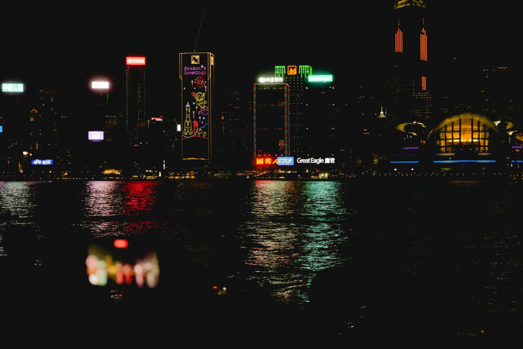 City skyline at night with brightly lit skyscrapers and colorful reflections on the water captures the essence of a weekend in Hong Kong. A person snapping a photo stands in the foreground, slightly blurred, adding to the bustling atmosphere of this vibrant city.
