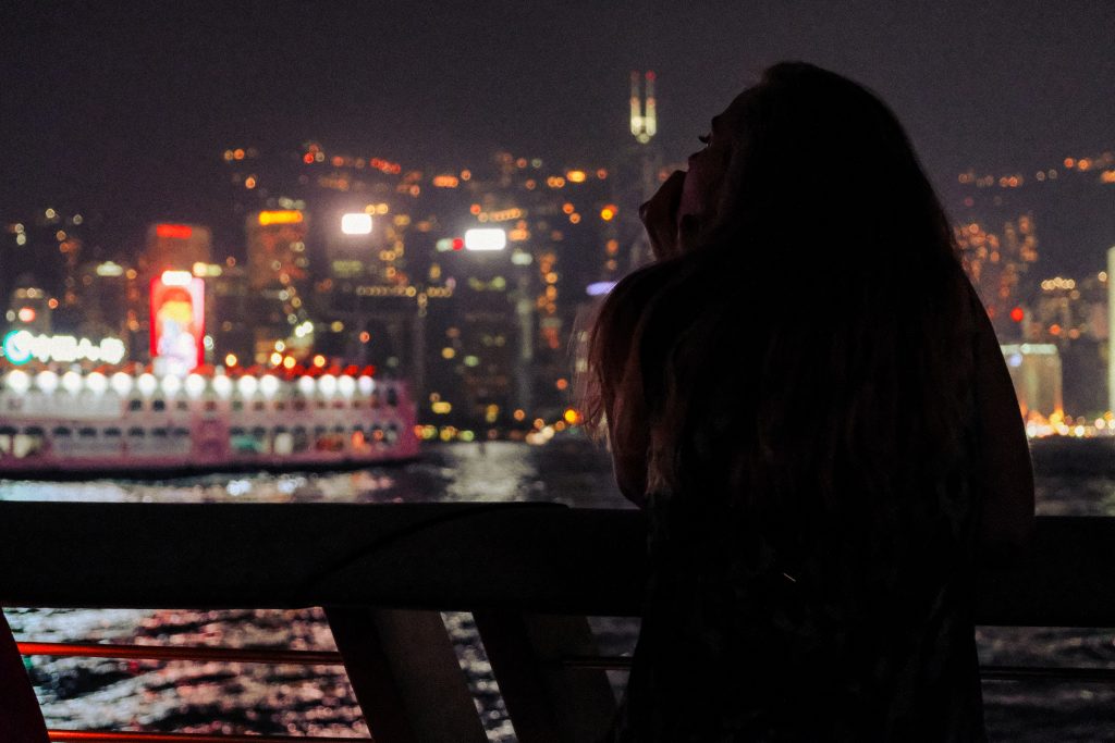 Sofia gazes out over the Hong Kong waterway at on a weekend night as the brightly lit ferry passes the city skyscrapers behind.
