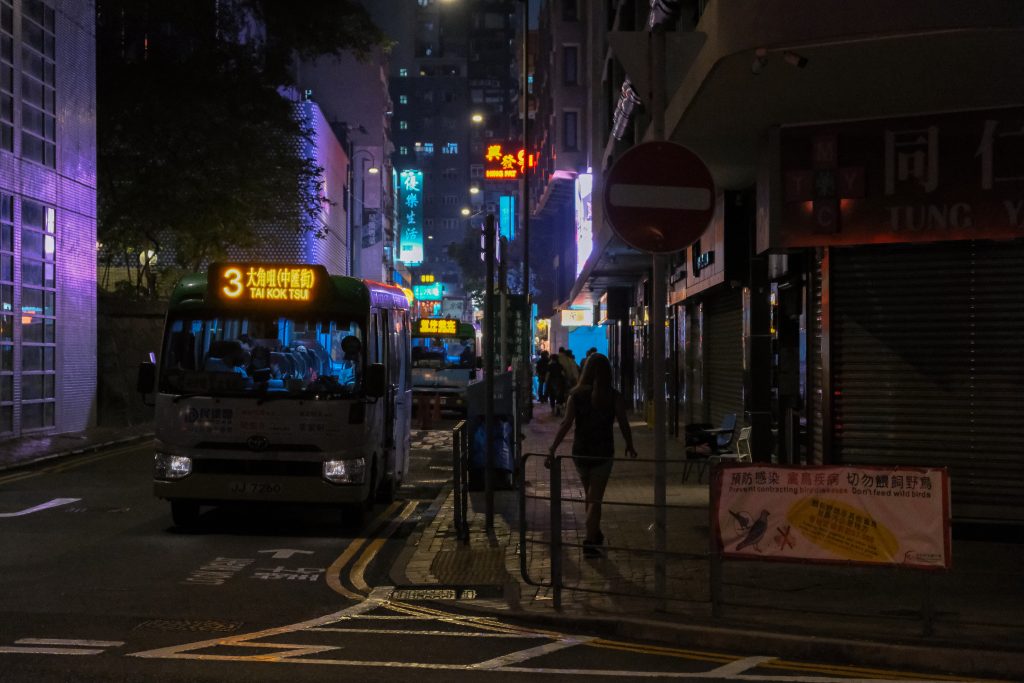 Bus on dimly lit urban street at night, with streetlights and neon signs illuminating the scene.