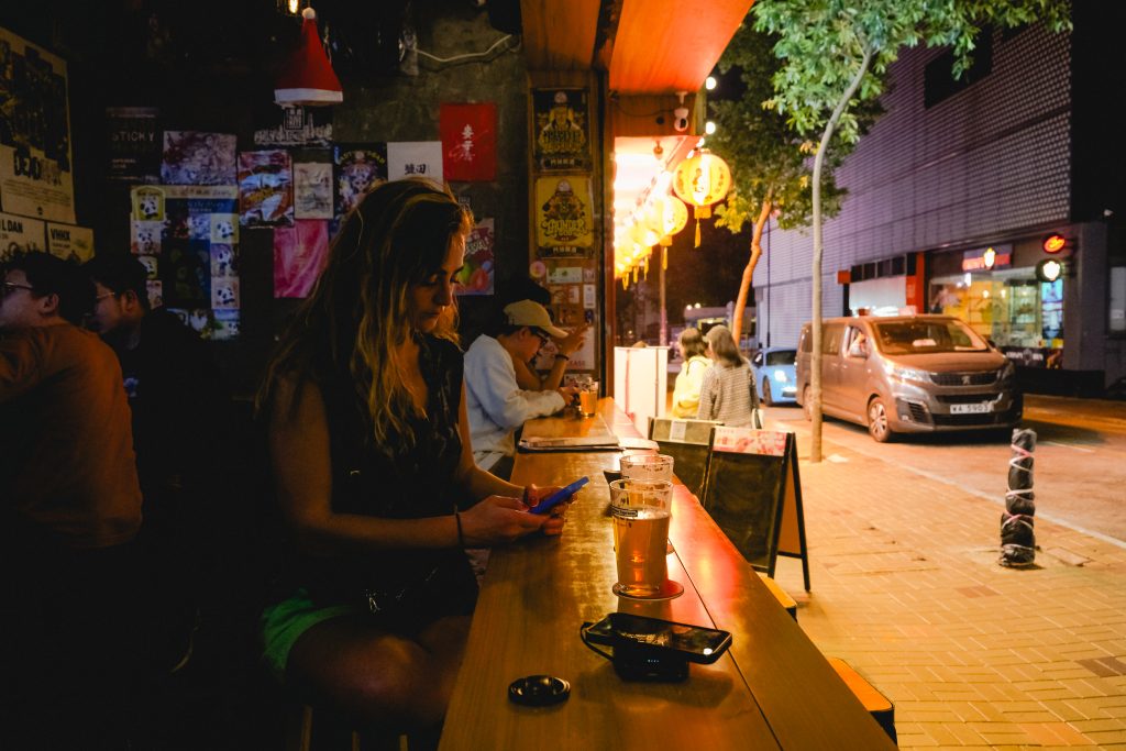 A woman using her phone sits at a dimly lit bar, with a drink and an ashtray on the counter. Street view outside.