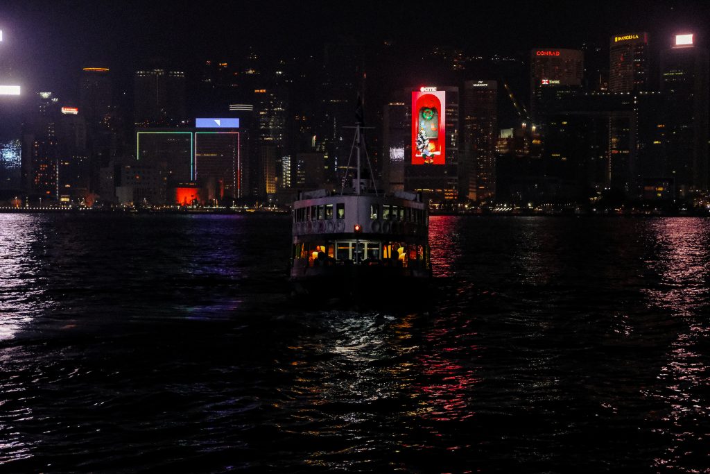 Ferry sailing towards a brightly lit city skyline at night, with colorful reflections on the water.