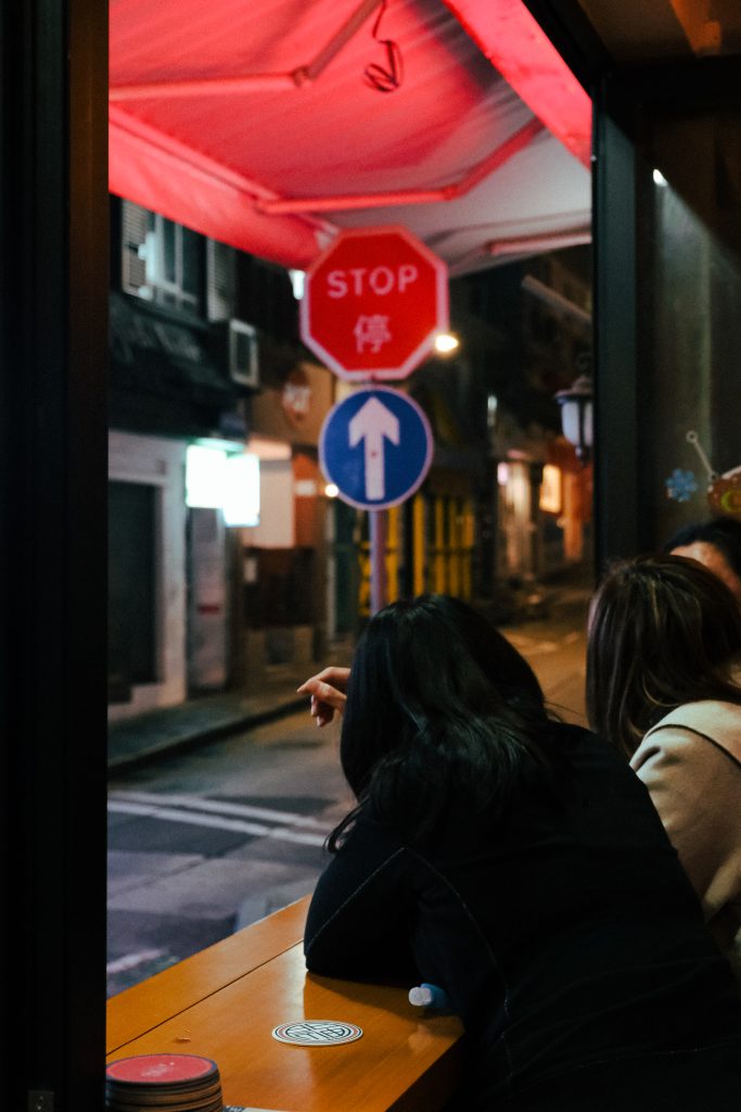 Two people sit by a window at night, with stop and direction signs outside on an empty street.