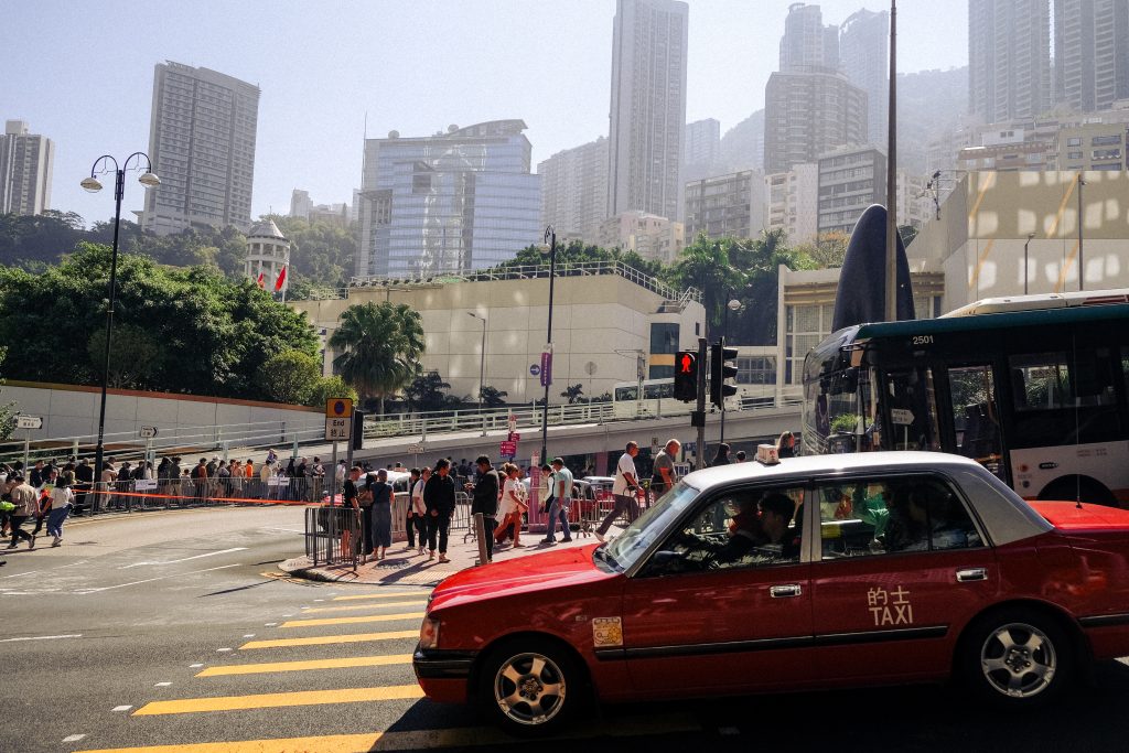 A red taxi waits at a yellow painted crosswalk as busy weekend crowds fill Hong Kong.