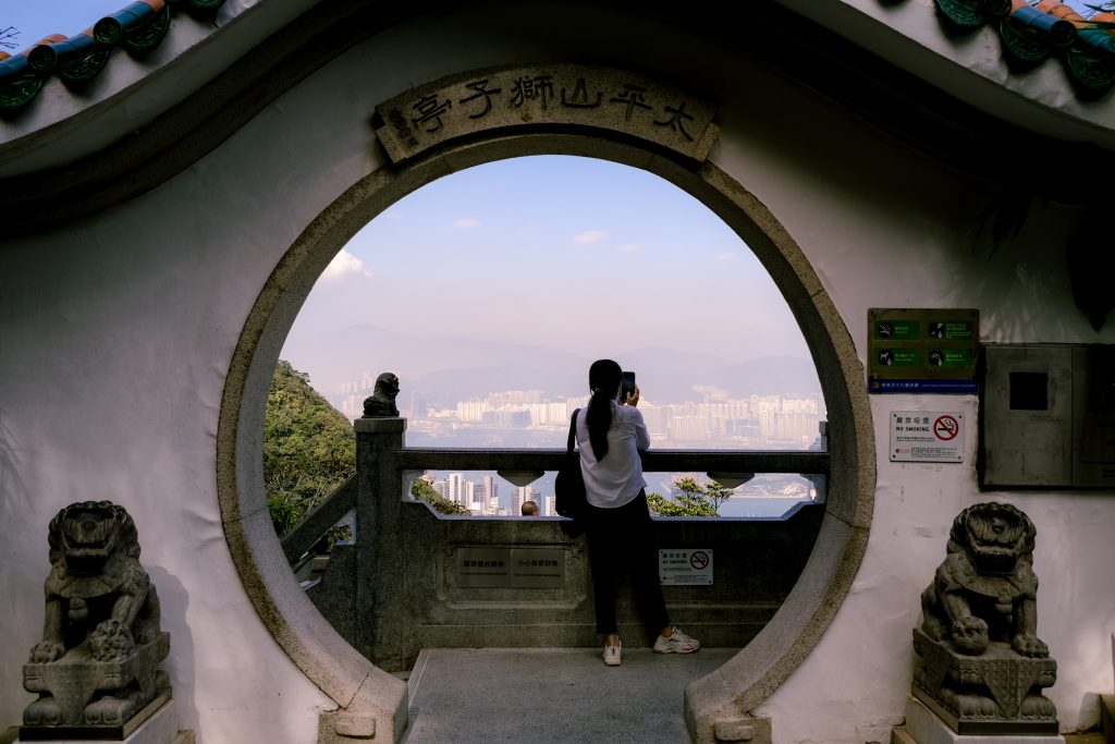 Woman taking a photo through a circular archway overlooking a cityscape, flanked by stone lion statues.