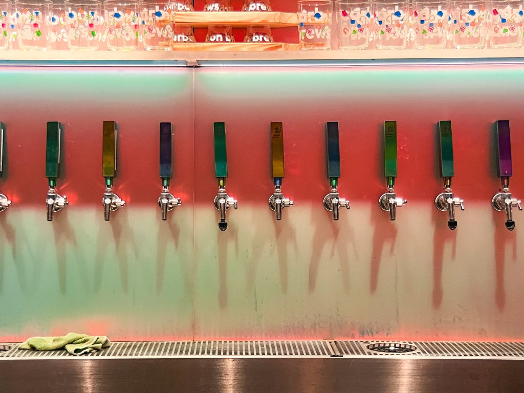 A row of colorful beer taps on a bar wall with a reflective countertop and a cleaning cloth.