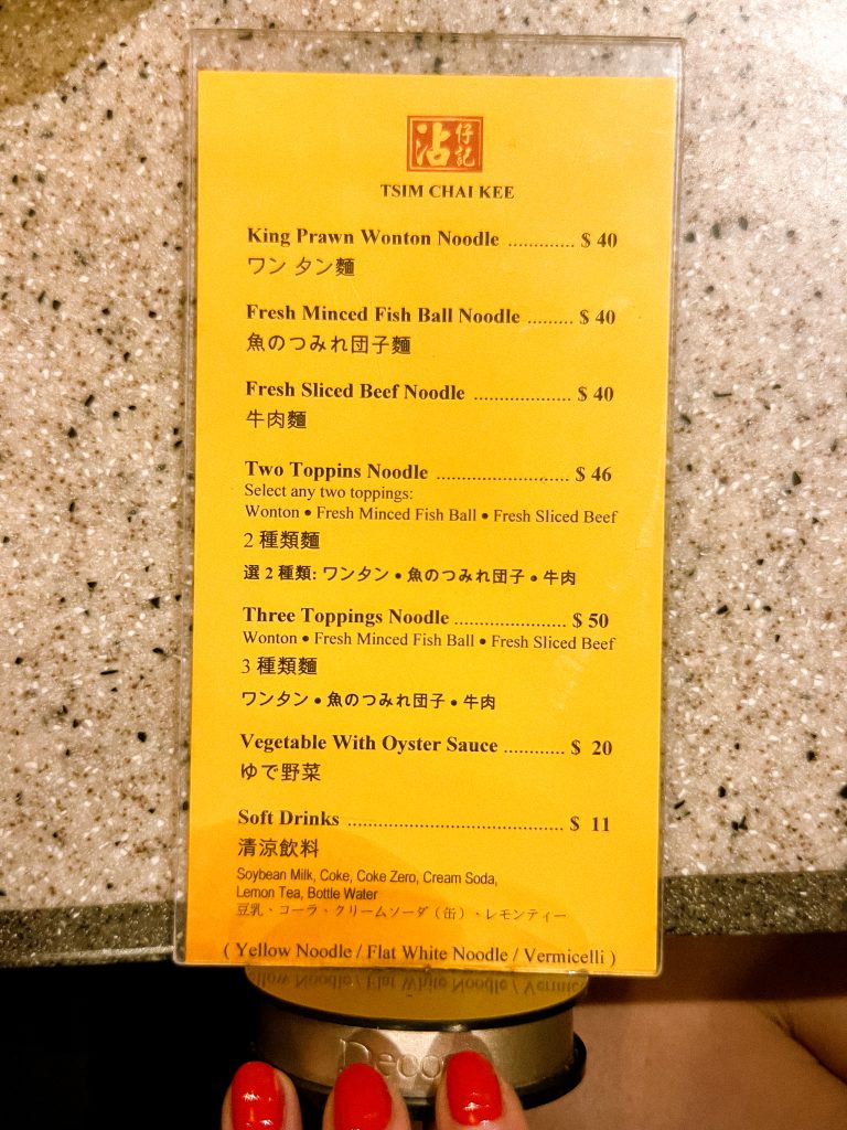 Menu on a table showing various noodle dishes with prices and brief descriptions, featuring English and Chinese text.