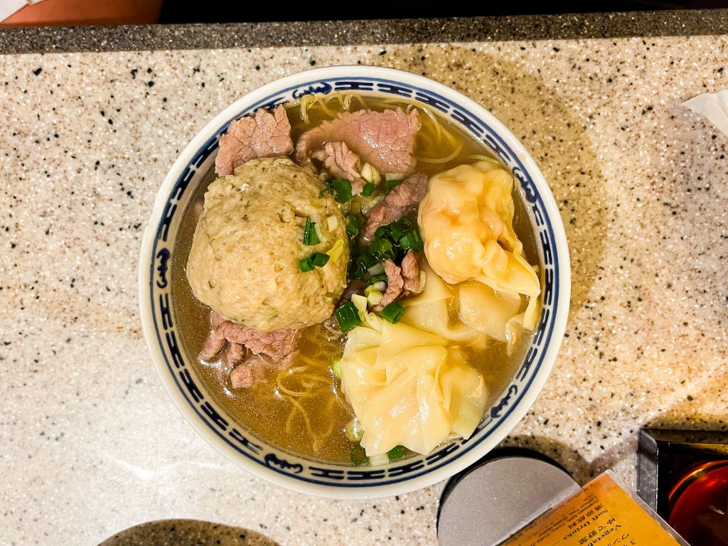 Bowl of soup with beef slices, a large meatball, wontons, and green onions on a speckled countertop.