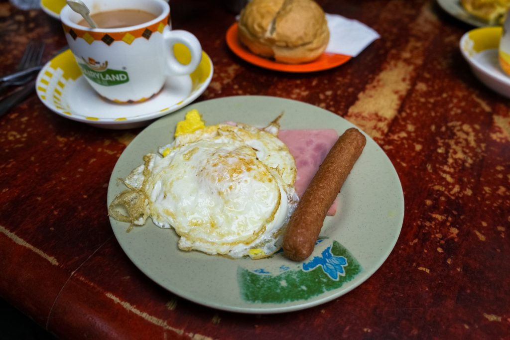 Plate with fried eggs, sausage, and ham. Cup of tea and a bun on a colourful table.