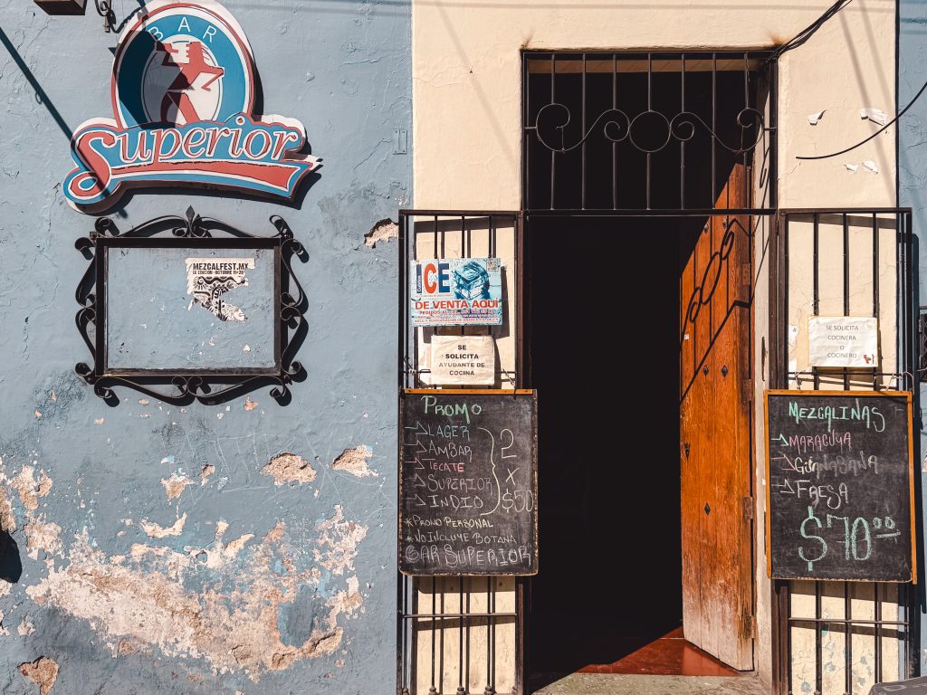 Entrance to a bar with a Bar Superior sign, chalkboards with promotions, and an open wooden door.