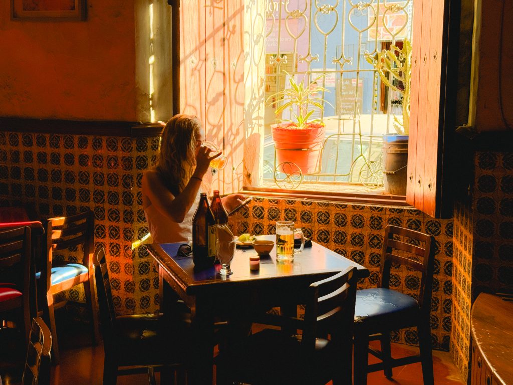 Sofia enjoying a drink at a sunlit table near a patterned wall and window with a potted plant.